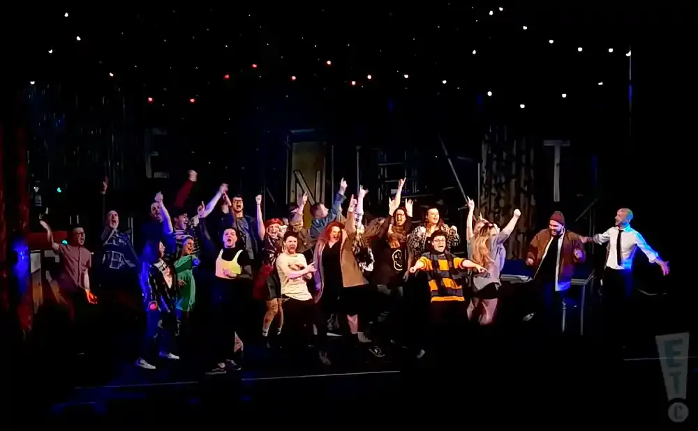 A diverse group of people on stage with their arms raised in celebration, with a dark background and small lights.