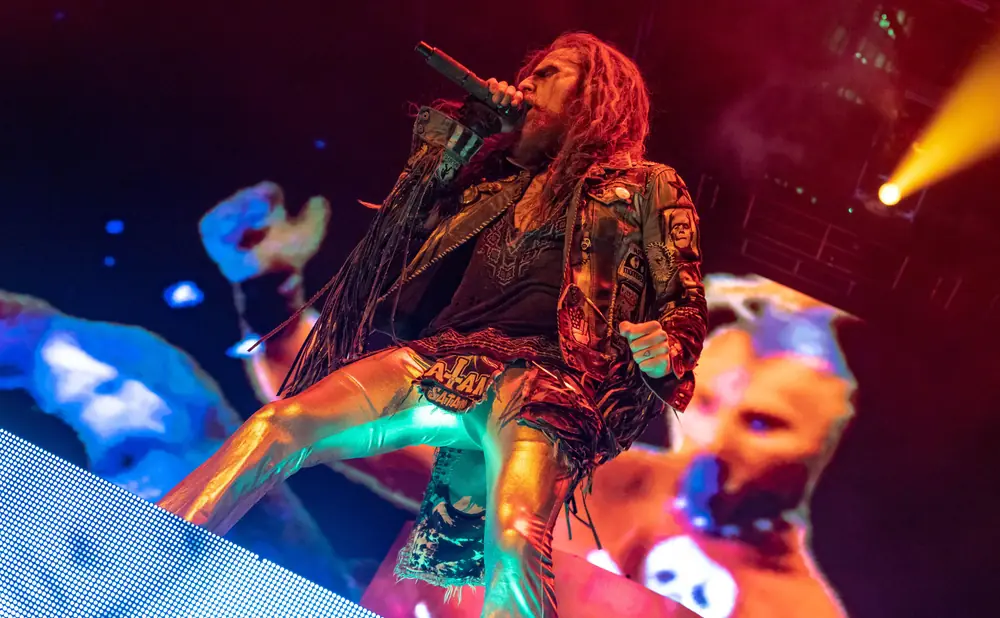 Rob Zombie performs on stage in a leather jacket with patches, metallic pants, and face paint.