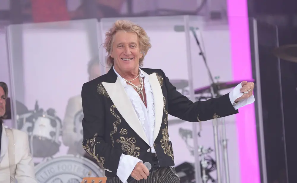 Rod Stewart performs on stage in a black sequined jacket and striped pants, wearing multiple necklaces.