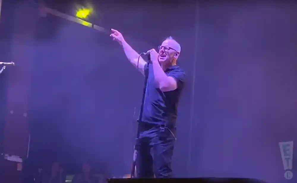 Singer with mohawk and glasses on stage, pointing and holding a microphone, under purple stage lights.