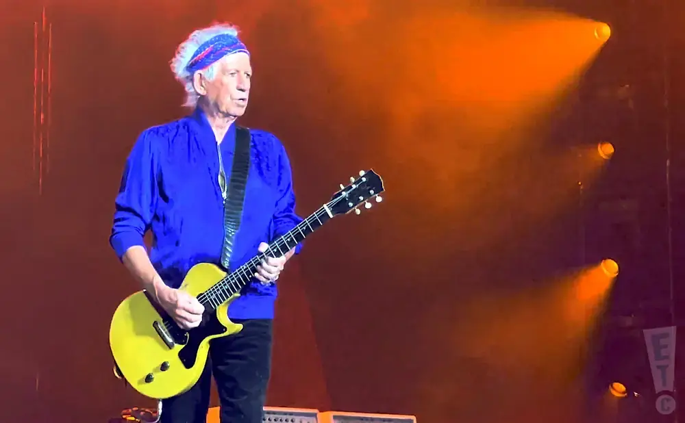 Keith Richards plays a yellow electric guitar on stage, bathed in orange spotlight.
