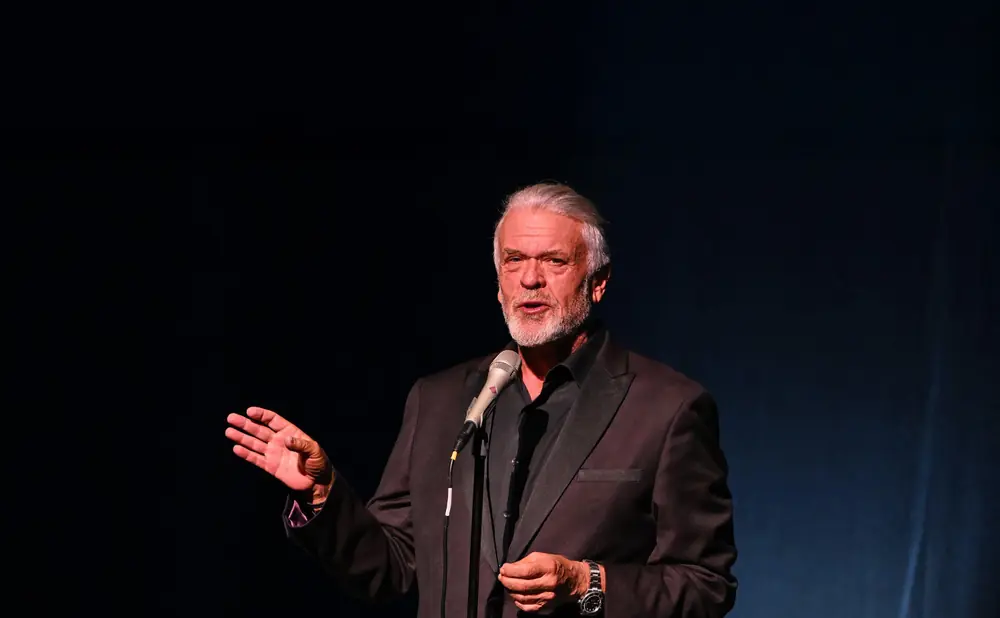 Man in a tuxedo speaks into a microphone, gesturing with his left hand.