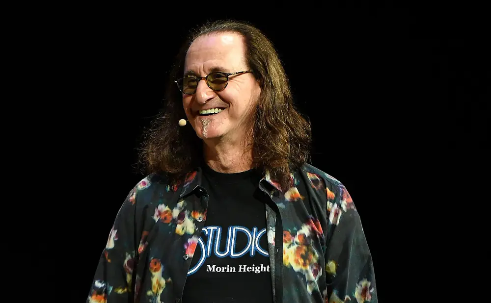 Geddy Lee of Rush smiles, wearing a floral shirt over a t-shirt that says 
