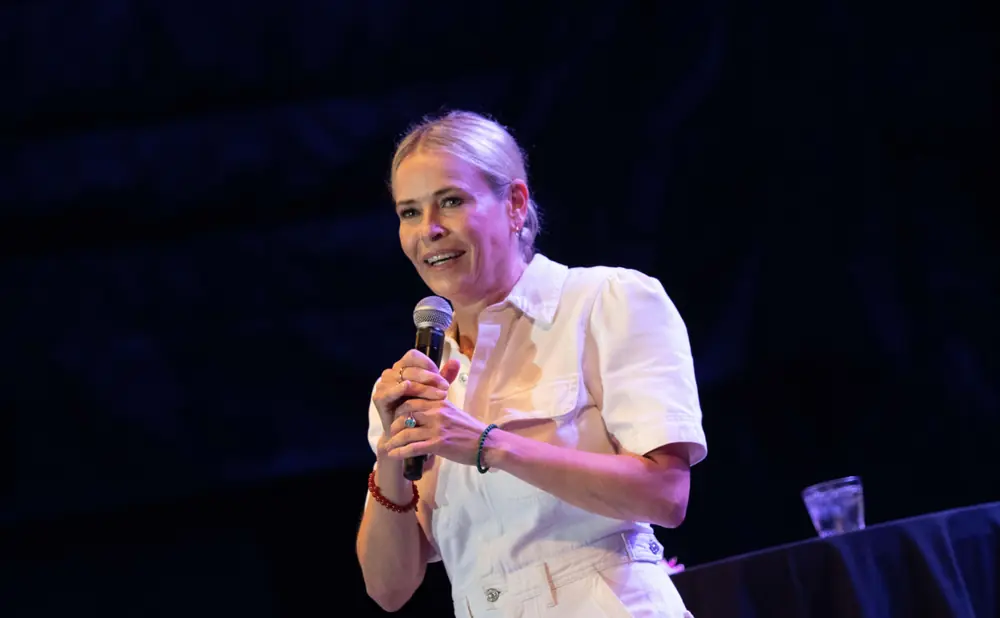 A woman with blonde hair smiles as she holds a microphone on a dark stage.