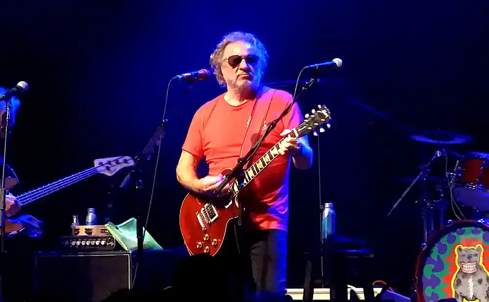 A man in a red shirt plays a red electric guitar on stage, illuminated by blue lights.
