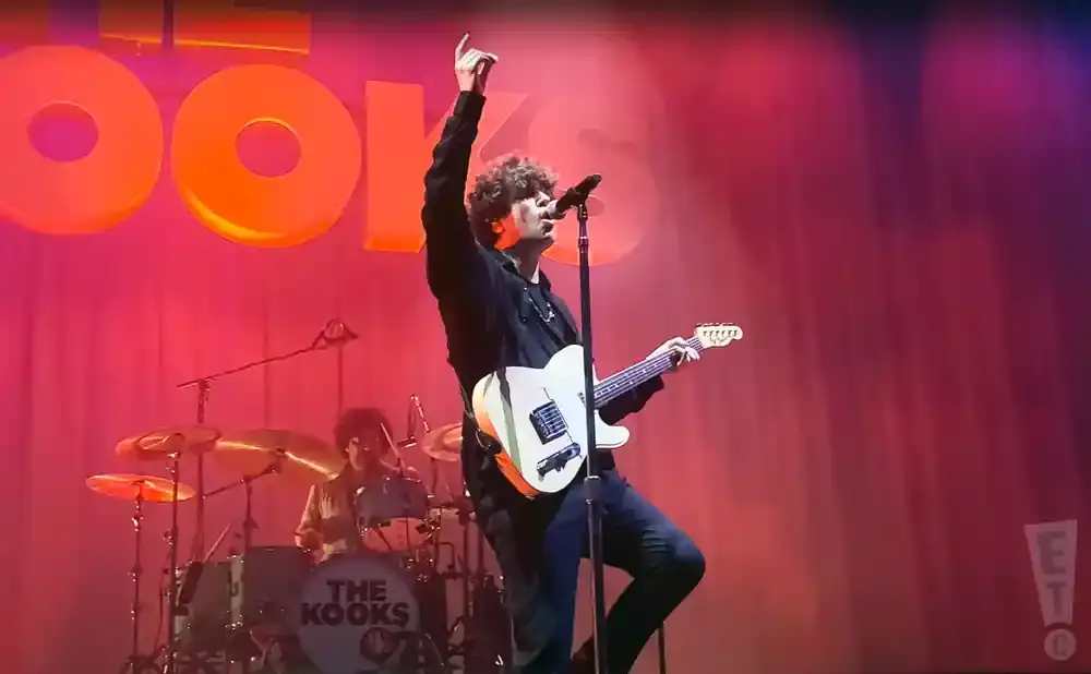 Singer of The Kooks performs on stage, raising one hand while playing a white electric guitar.