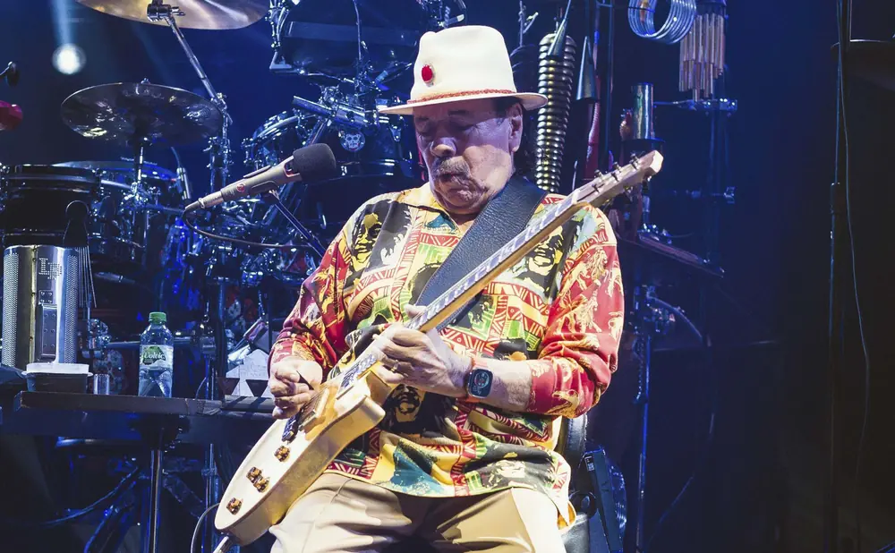 Carlos Santana plays a white guitar on stage, wearing a colorful shirt and white hat.