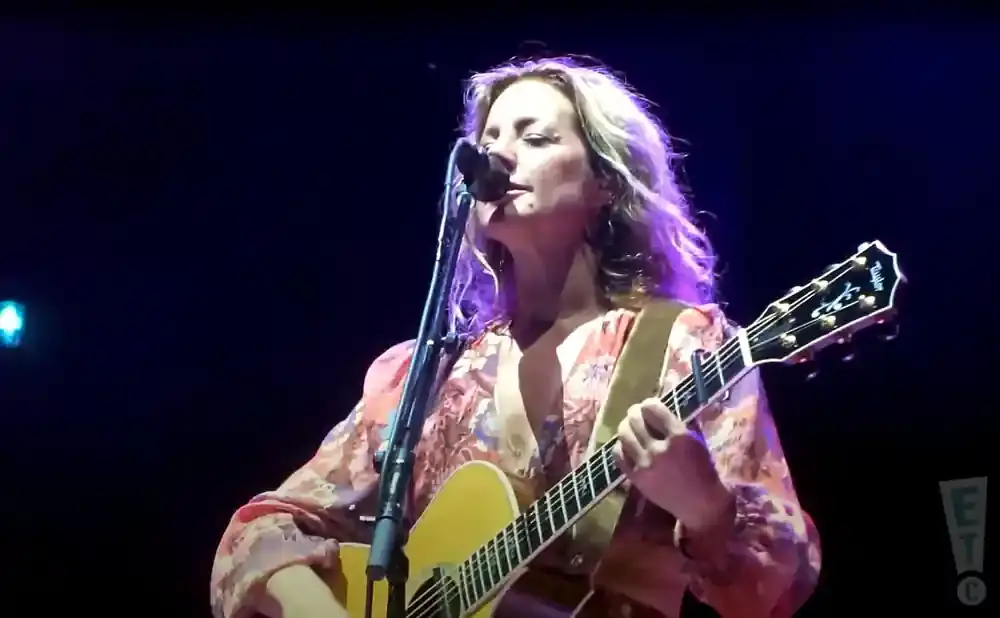 A woman with blonde, wavy hair sings into a microphone while playing an acoustic guitar.