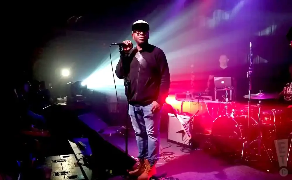 A man in a black jacket, jeans, and a white cap sings into a microphone on a dimly lit stage with purple and pink lights.