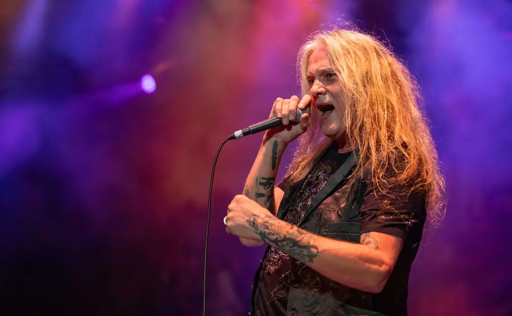 A male singer with long blonde hair, tattoos, and a sparkly vest sings into a microphone on a dimly lit stage.