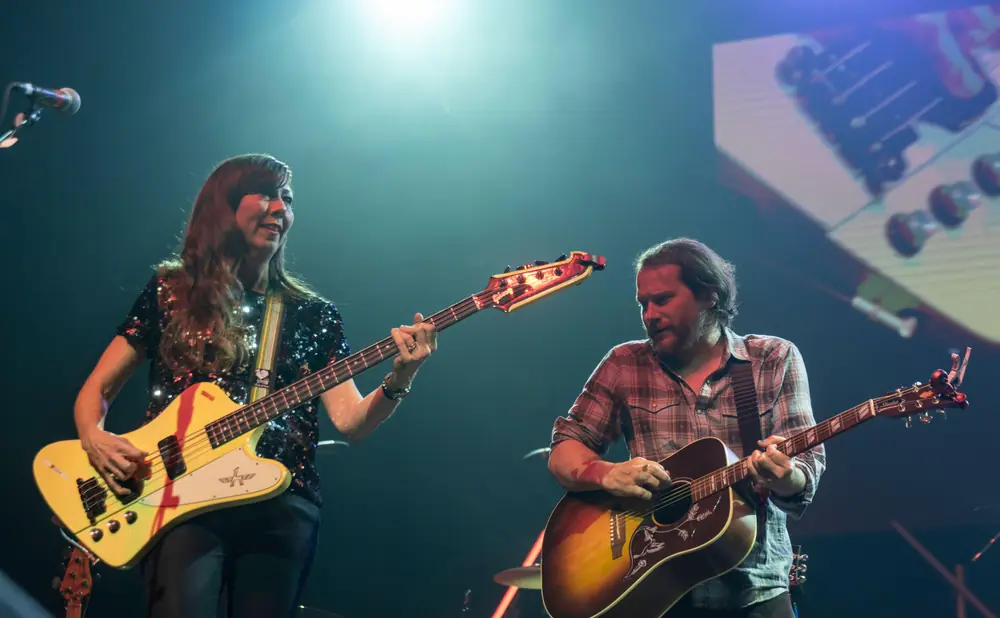 Two musicians play guitars on stage, bathed in blue and white stage lights.