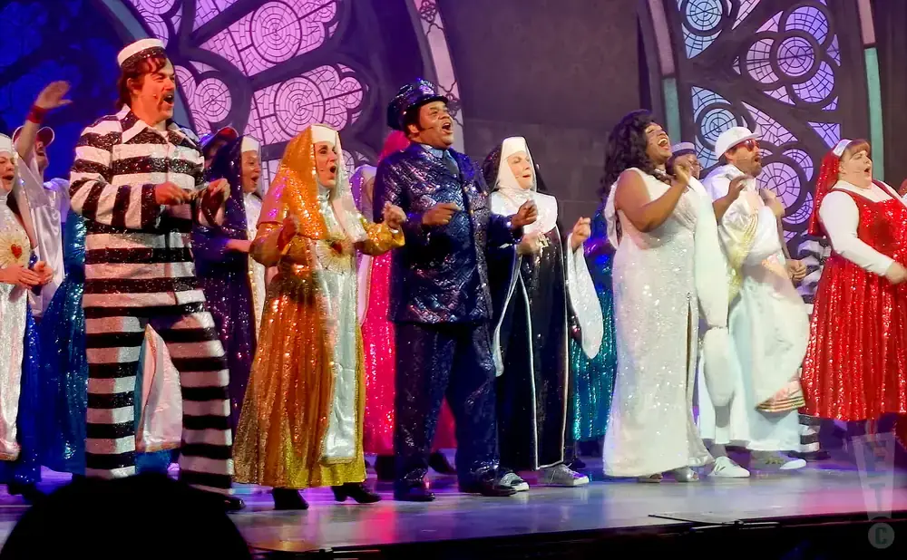 A group of performers in glittering costumes, including a prisoner in black and white stripes and several in sequined nun habits, stand on stage.