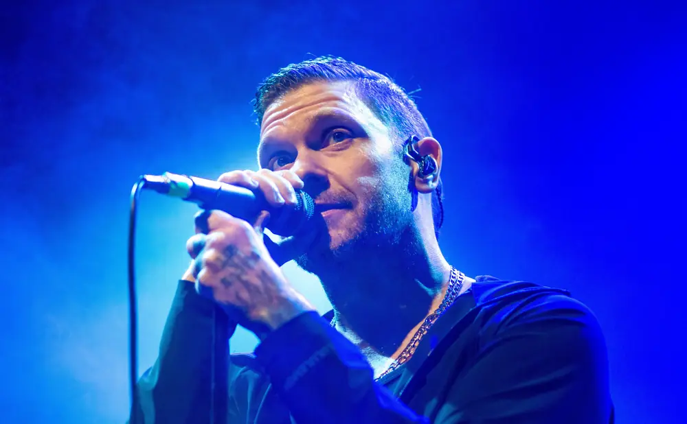 A man with tattoos on his hands sings into a microphone on a blue-lit stage.