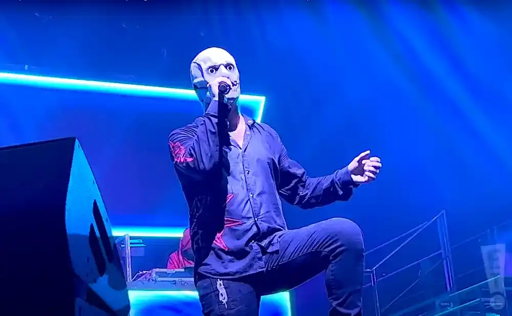 A performer in a white, mask-like helmet sings into a microphone on a stage bathed in blue light.
