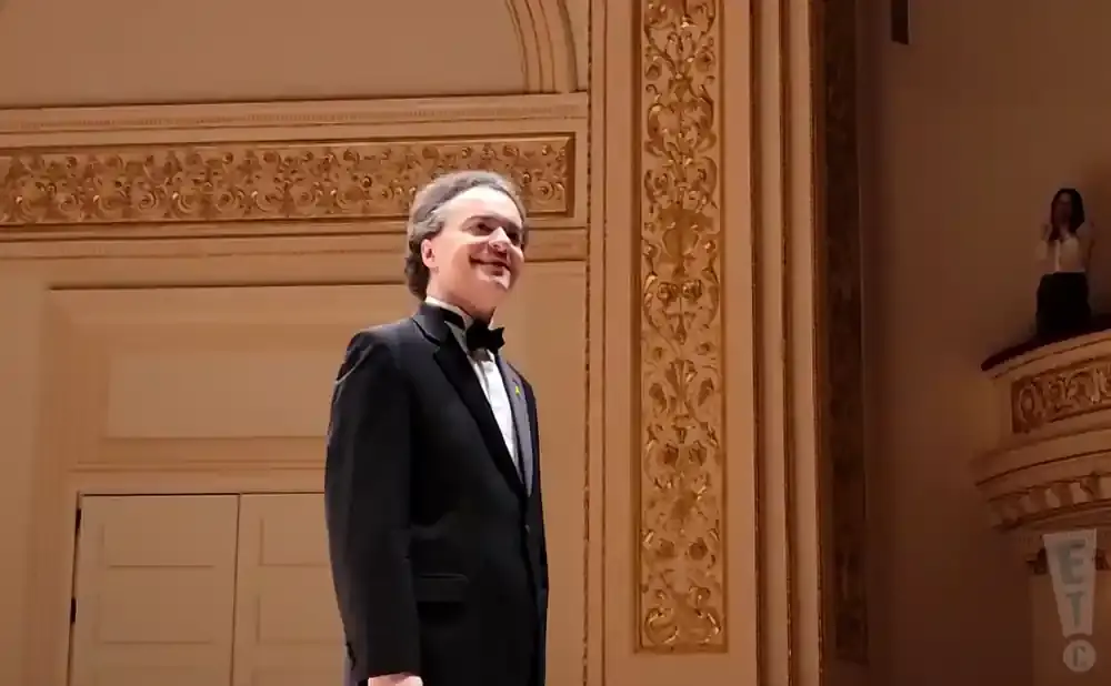 A man in a tuxedo smiles on a stage with ornate gold decor.