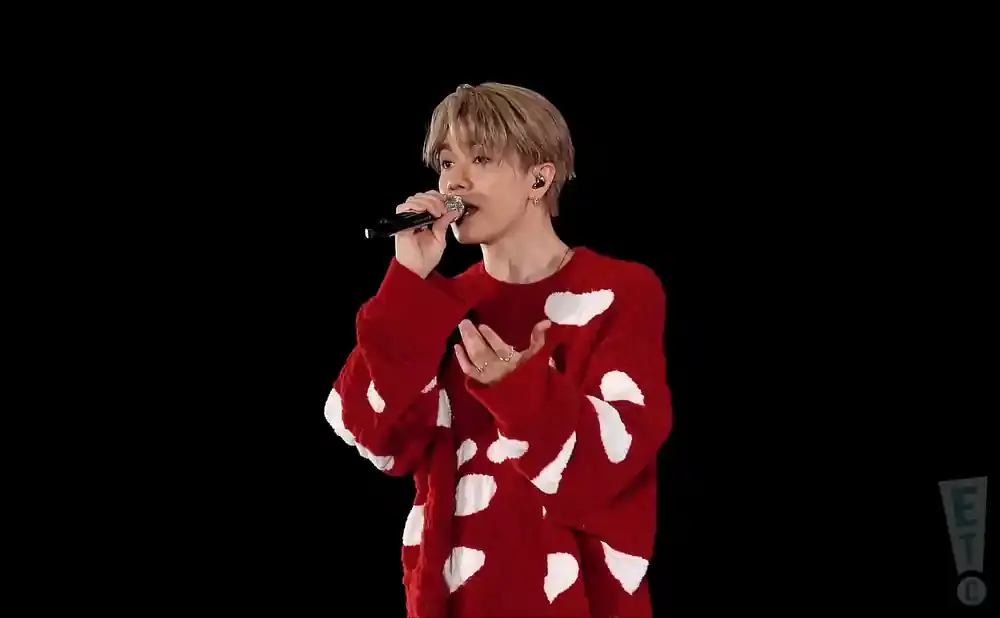 A man with blonde hair sings into a microphone wearing a red sweater with white cloud-like patterns.