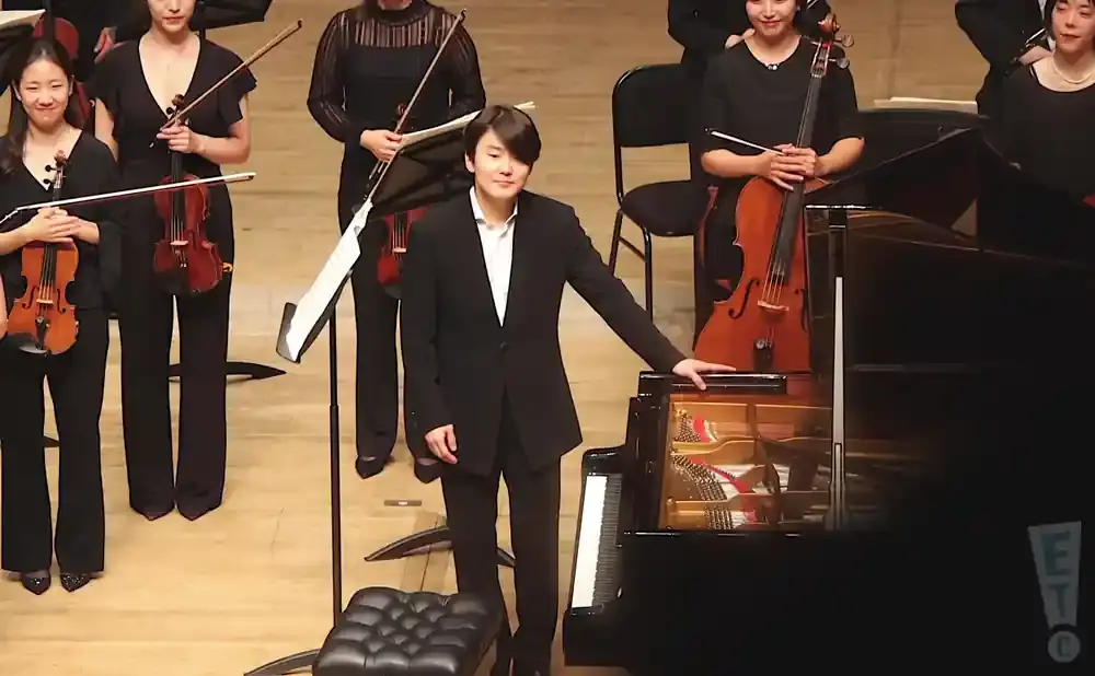 Pianist in a black suit stands by a grand piano, with an orchestra behind him.