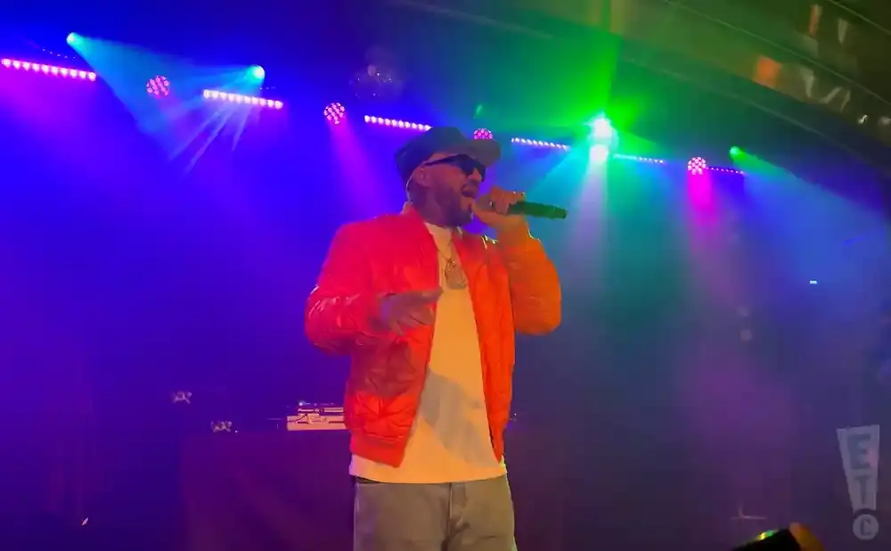 A man in an orange jacket and baseball cap sings into a microphone on a stage lit by purple and green lights.