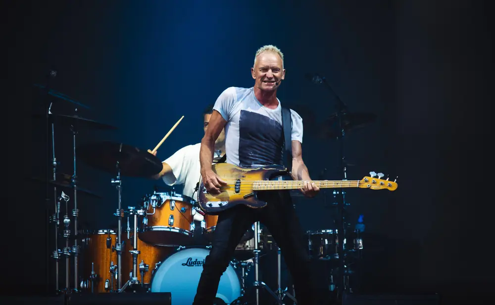 Bassist Sting smiles while playing a Fender bass guitar during a live performance, with a drummer in the background.