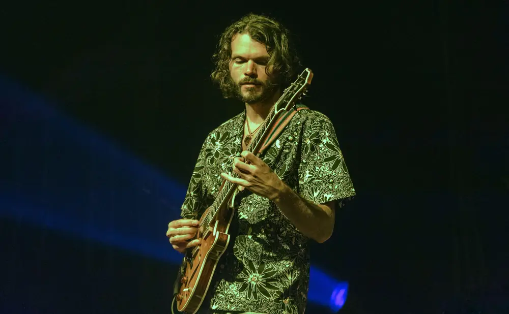A man with long, dark, curly hair and a beard plays a guitar on stage, lit by blue and green stage lights.
