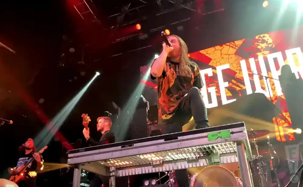 A male singer with long hair holds a microphone while performing on stage with a band.