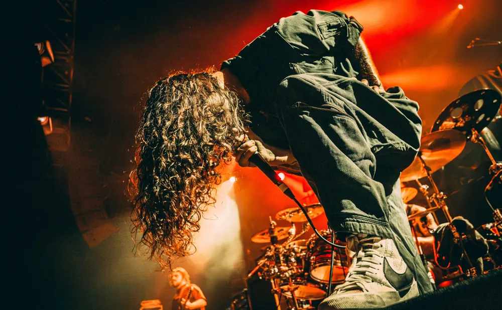 A rock singer with long, curly hair leans into a microphone on a stage lit with red and orange lights.