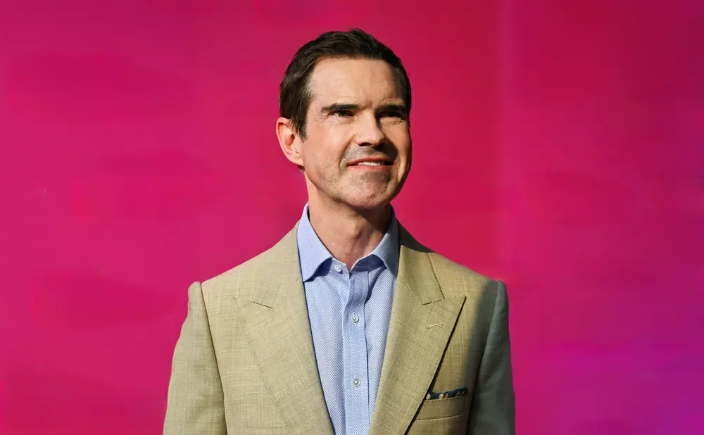 Man in a tan suit and blue shirt smiles against a bright pink background.
