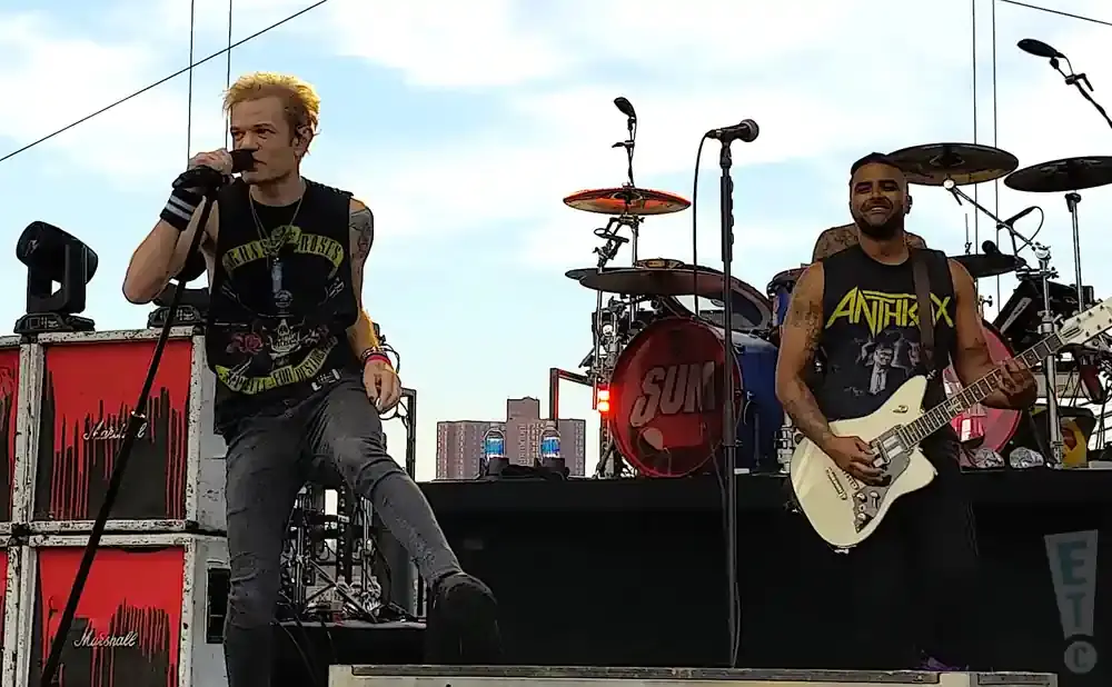 Two men perform on stage with guitars and drums, one singing into a microphone.