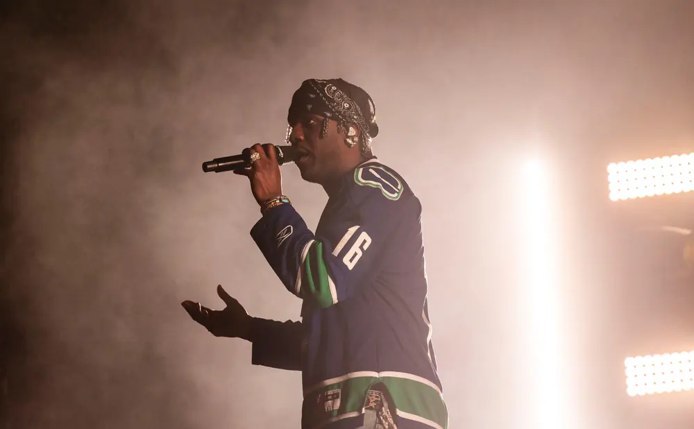 A rapper performs on stage wearing a blue and green hockey jersey, bandana, and jewelry.