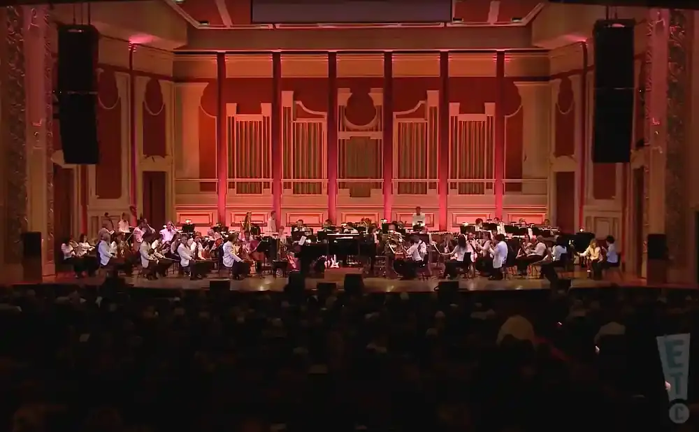 An orchestra performs on stage in front of a large audience in a grand hall.