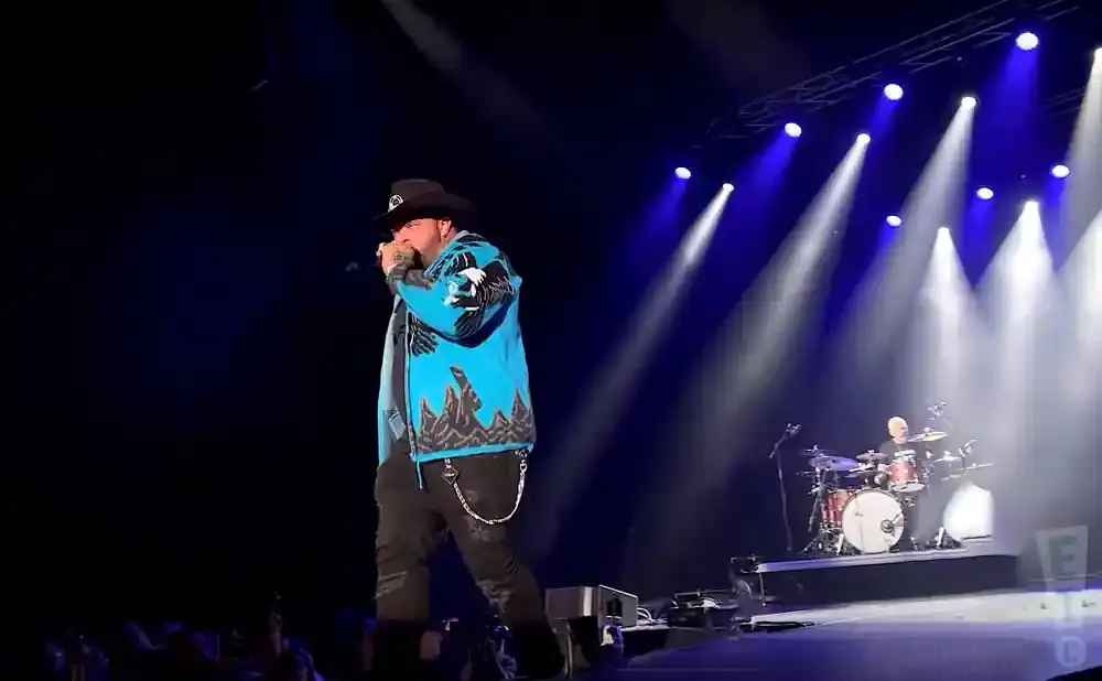 A male singer in a cowboy hat and bright blue jacket performs on stage with a drummer in the background.