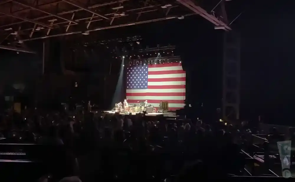 A band performs on stage in front of a large American flag backdrop at an indoor concert.