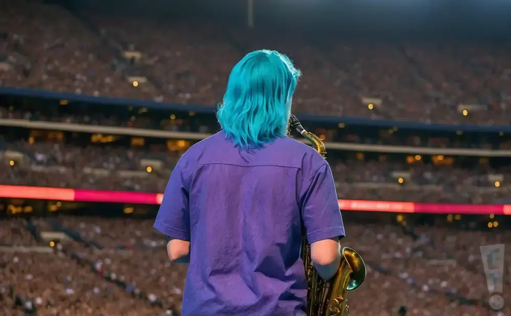 A person with bright blue hair plays a saxophone on a stage in front of a stadium full of people.