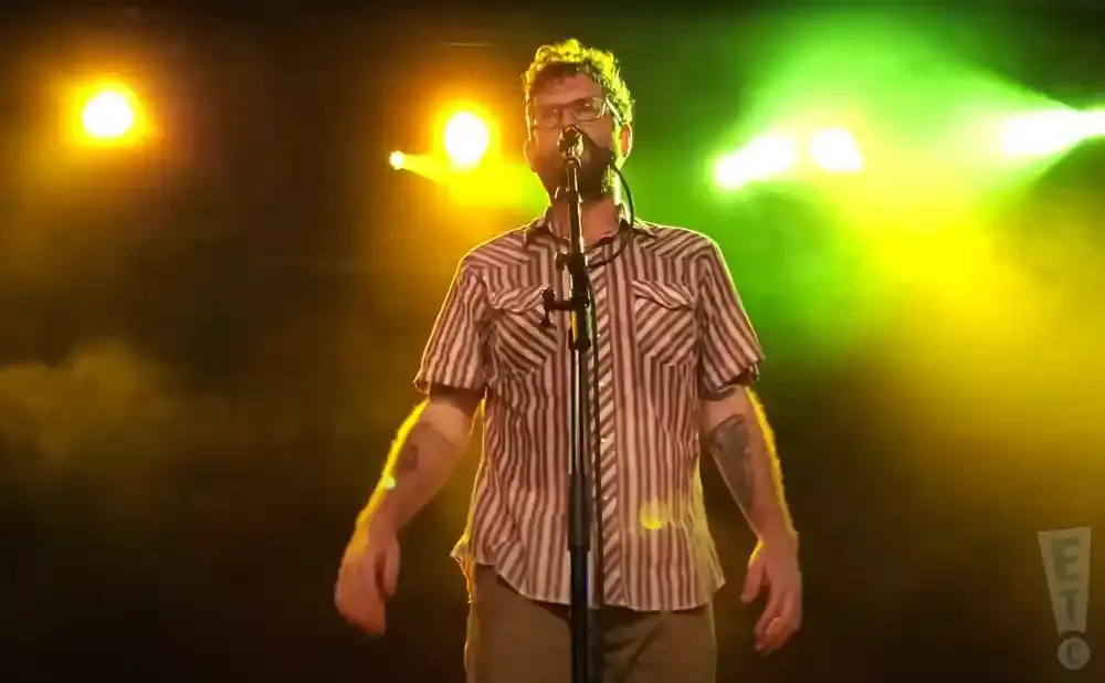 A man with glasses and a striped shirt stands on stage, singing into a microphone.