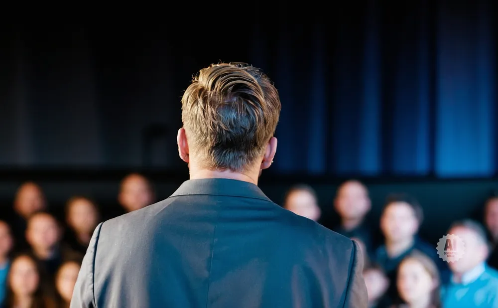 Man in suit facing away from camera, addressing a blurry audience.
