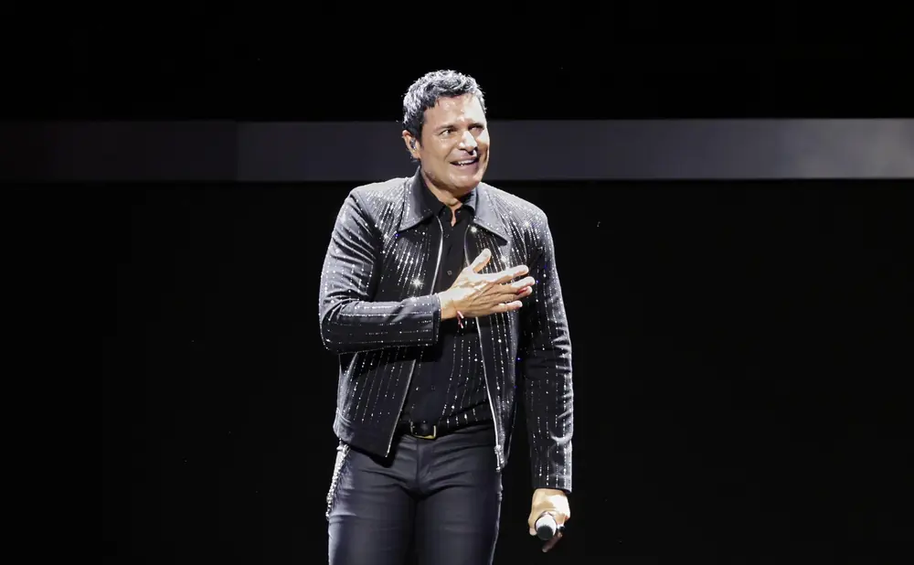 Singer Chayanne smiles and places his hand on his chest on stage, wearing a sparkling black jacket.