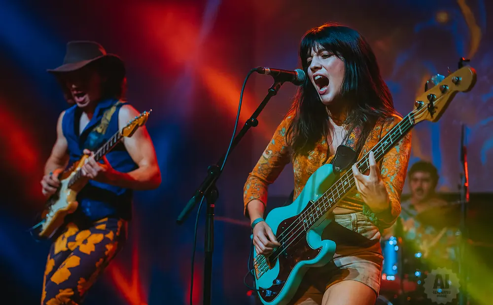 A woman sings and plays a bass guitar on stage, with a man playing guitar beside her.