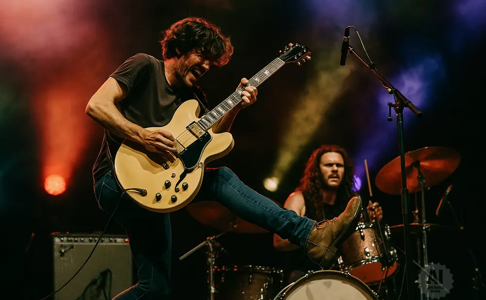 A guitarist performs on a cream-colored electric guitar, with a drummer in the background.