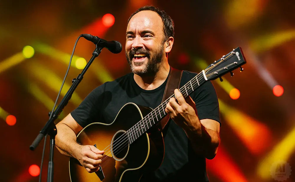 Dave Matthews smiles while playing his guitar on stage with colorful lights in the background.