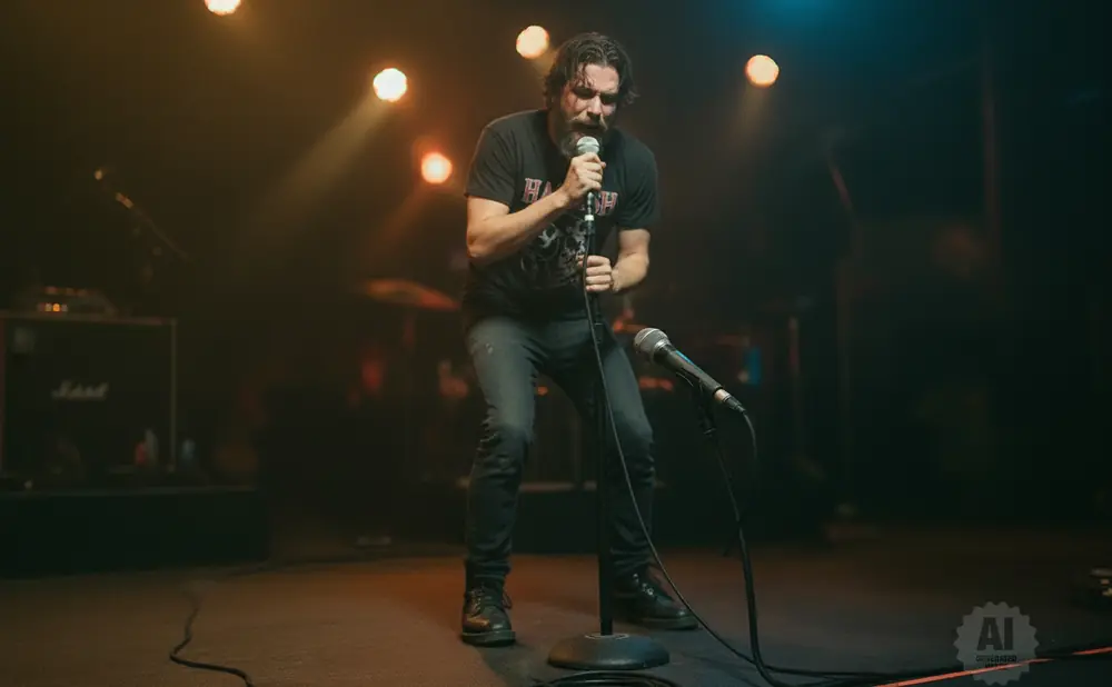 A male singer performs into a microphone on a dark stage, lit by warm spotlights.