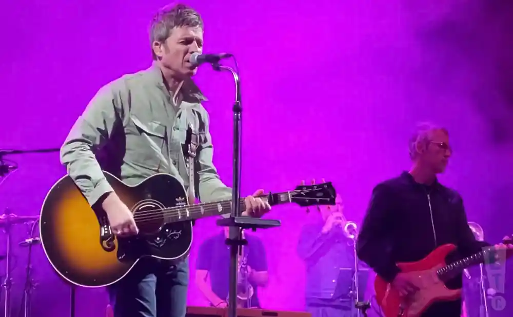 noel gallagher performing live on stage playing the guitar in front of purple light