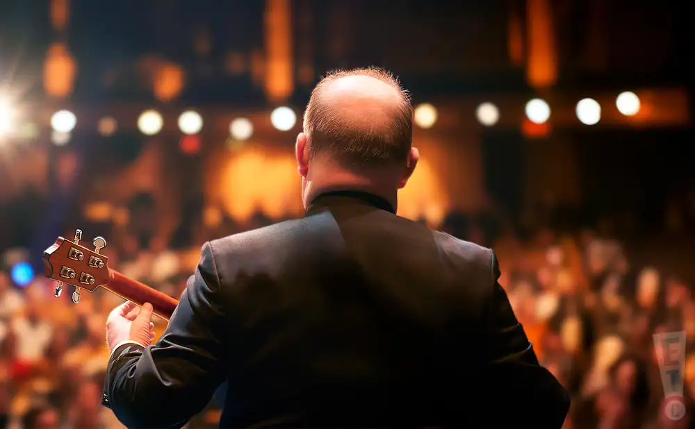 a rear view photo of george hinchcliffes ukulele orchestra of great britain performing a concert on stage to a lively audience