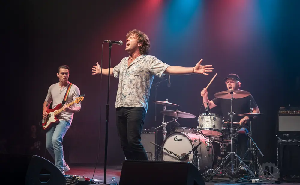 A band performs on stage with a singer in the center, a guitarist on the left, and a drummer on the right.