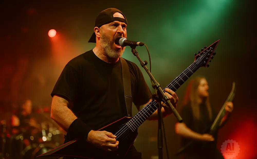 A man with a beard and a baseball cap sings into a microphone while playing a black electric guitar on stage.
