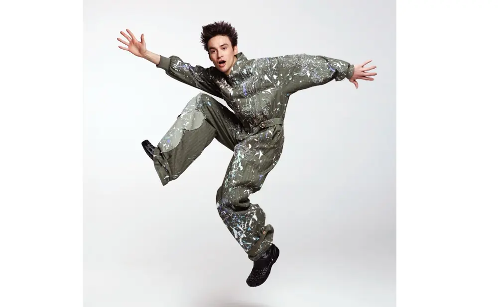 A person in a paint-splattered jumpsuit jumps with arms outstretched against a white background.