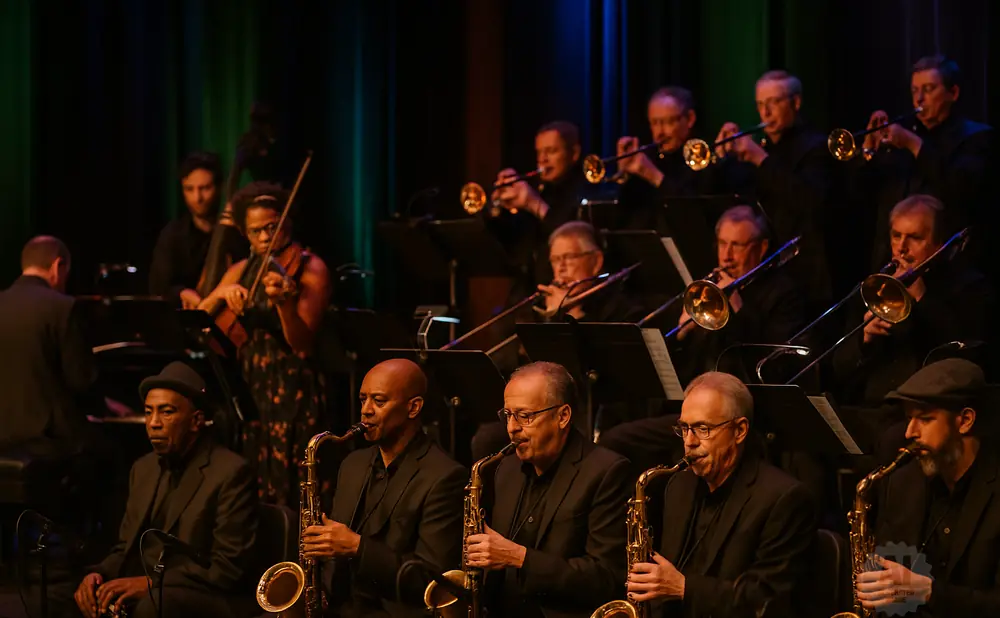 A jazz orchestra performs on stage, with musicians playing brass instruments and a violinist.