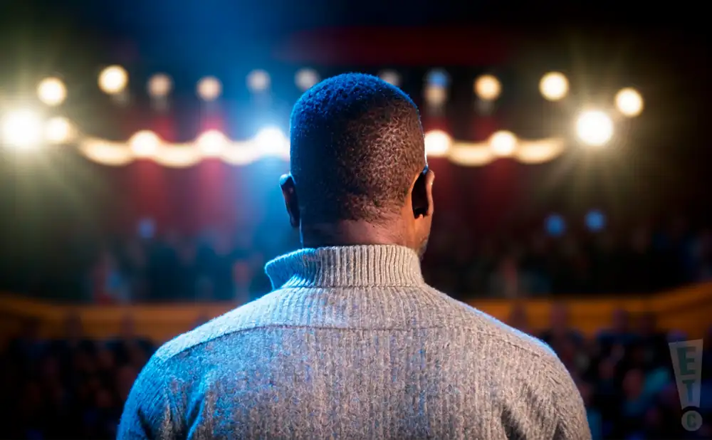 a rear-view photograph from behind saxophonist merlon devine, performing on stage in front of a large audience.