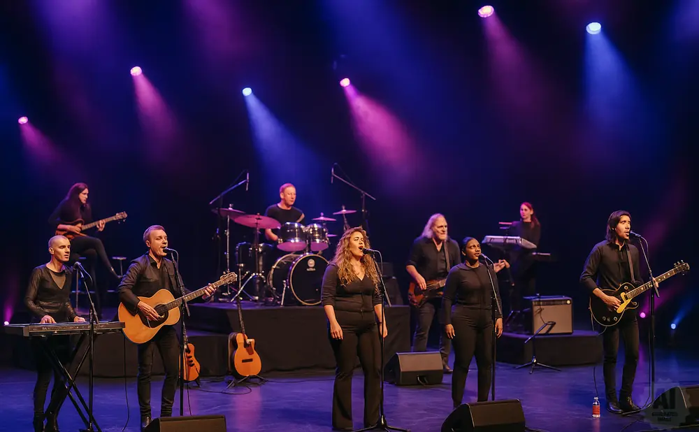 A band performs on stage with purple and blue lights. The band members play guitars, drums, and keyboards.