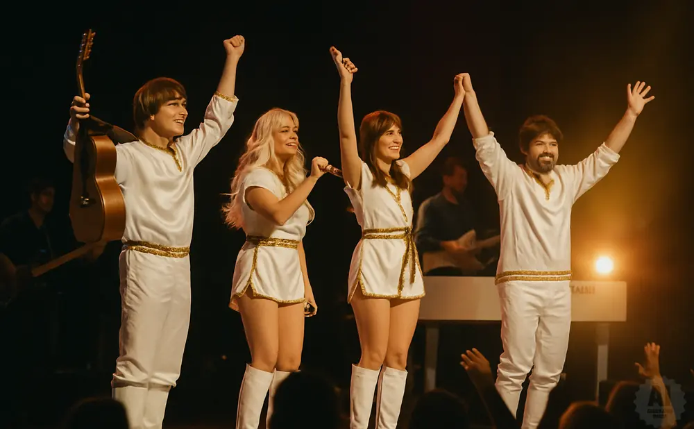 Four performers in white, gold-trimmed costumes, two women singing, two men playing instruments on stage.