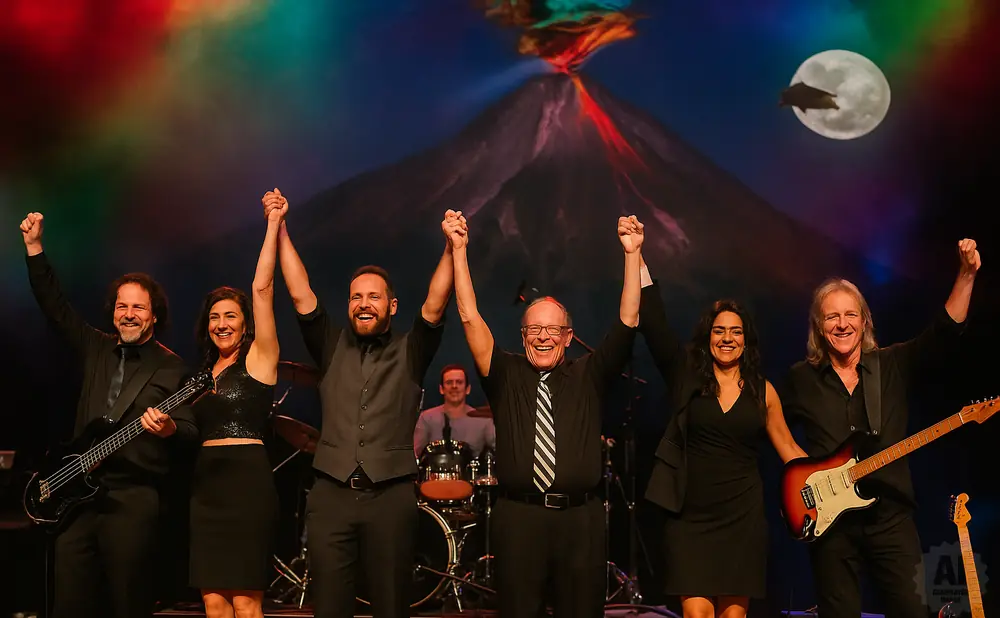 A band of six people, dressed in black, pose with their hands raised in celebration on stage with a volcano backdrop.